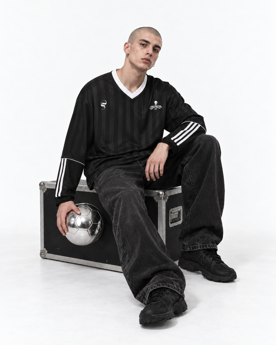 A man sits on a black equipment case wearing a black Adidas soccer jersey with white stripes, black jeans, and black sneakers, holding a silver soccer ball against a white studio background.